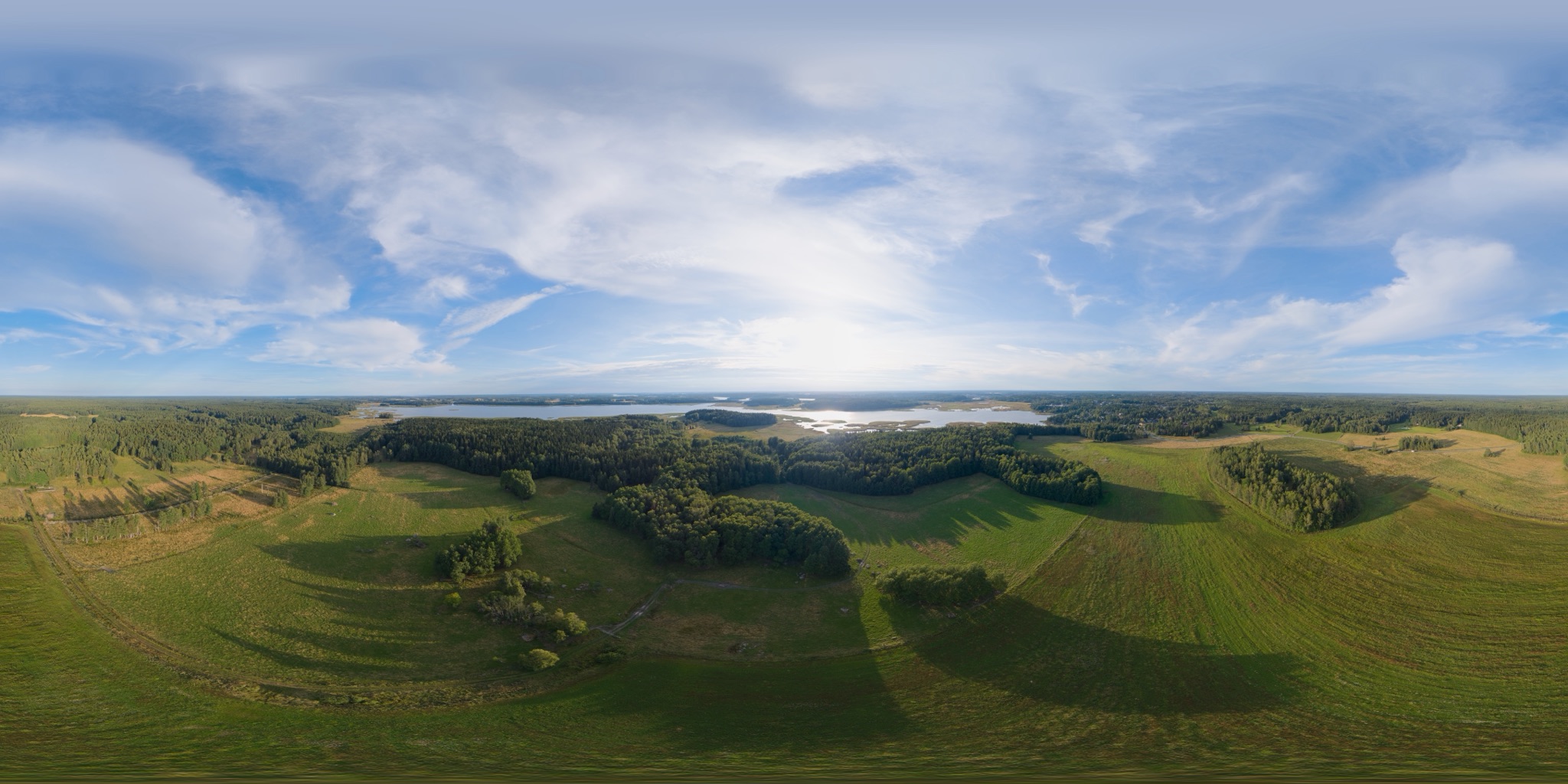 A 360-degree drone panorama laid out flat: green fields and patches of forest with long evening shadows, and a lake with reed islands towards the low sun under a blue sky with thin clouds.